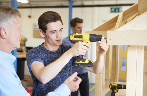 een mbo-student bedient een boormachine, naast hem staat een docent het te observeren en op de achtergrond is een andere mbo-student aan het werk