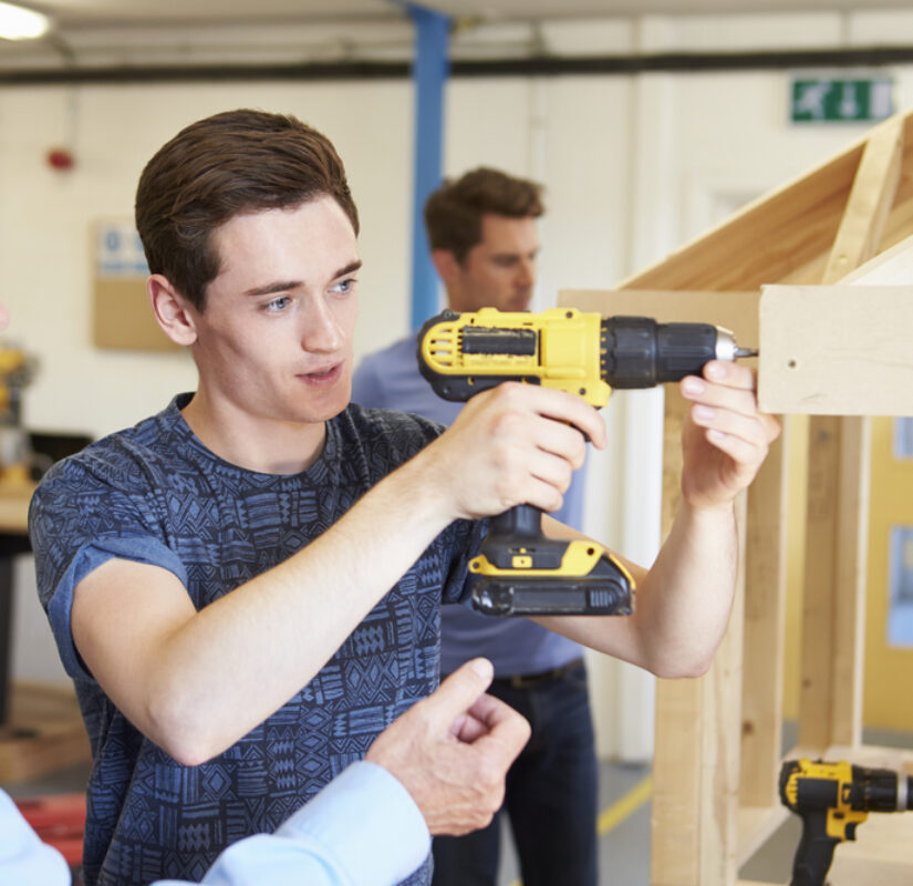 een mbo-student bedient een boormachine, naast hem staat een docent het te observeren en op de achtergrond is een andere mbo-student aan het werk
