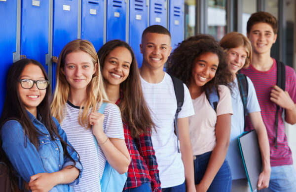 zeven scholieren staan naast elkaar glimlachend in de kamera, ze staan voor een muur met kluisjes in een schoolgebouw