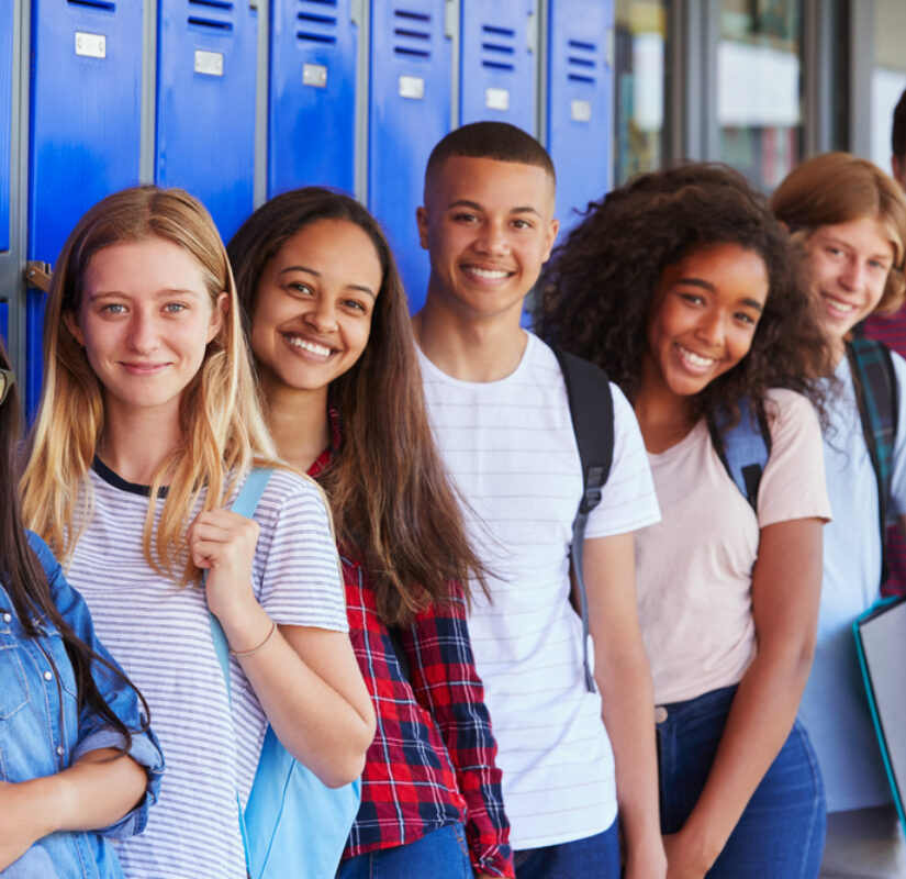 zeven scholieren staan naast elkaar glimlachend in de kamera, ze staan voor een muur met kluisjes in een schoolgebouw