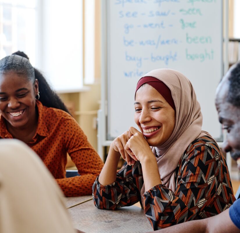 Drie mensen zitten in een klaslokaal naast elkaar, ze kijken heel blij, op de achtergrond zie je een schoolbord met daarop woorden